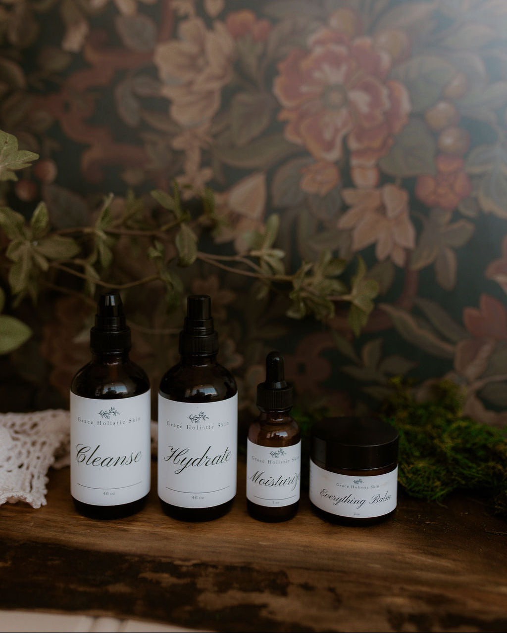 Set of skincare: Cleanse, Hydrate, Moisturize, Everything Balm in front of a dark floral wallpaper